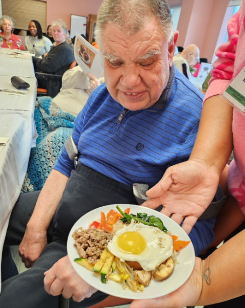 A person seated at a dining table is being served a plate of food. The plate contains a sunny-side-up egg on top of rice, surrounded by assorted vegetables, including carrots, spinach, zucchini, mushrooms, and a portion of ground meat. Other diners and tables with white tablecloths are visible in the background.