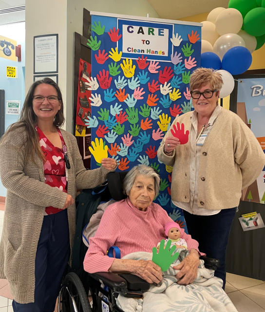 Three individuals pose for a photo in front of a hand hygiene awareness display. The display is covered with cut-out paper hands in various colours, with a sign at the top that reads “CARE TO Clean Hands.” An older adult is seated in a wheelchair with a blanket, holding a green paper hand and a baby doll. Two people stand beside the person in the wheelchair, holding up paper hands. The background includes balloons in white, blue, and green, and part of a bulletin board with additional decorations.