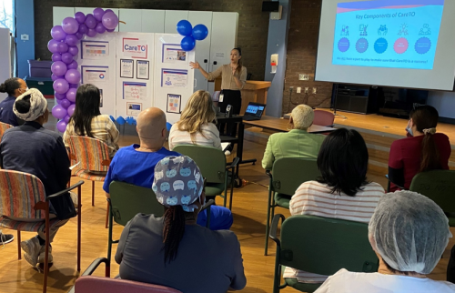 A person is giving a presentation to a seated audience in a room with wooden floors. The presenter stands near a display board decorated with purple balloons and informational posters labelled “CareTO.” Behind the presenter, a large screen shows a slide titled “Key Components of CareTO” with colourful icons and text. The audience is seated in rows of chairs, and some individuals are wearing medical scrubs and hairnets.