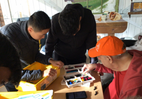 Several people are gathered around a table working on a hands-on activity with robotics kits. The table has open containers filled with colourful LEGO pieces and components, and a smartphone is placed nearby. One person wearing an orange cap is pointing at pieces in the tray, while others lean in to examine and select parts.