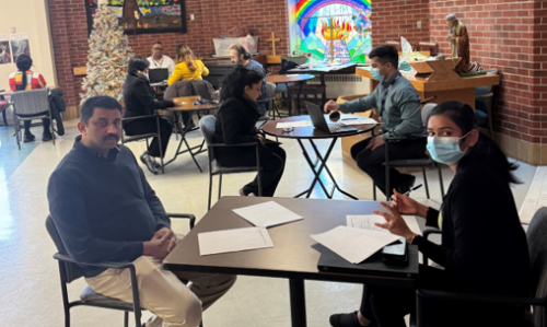Several pairs of people are seated at small round and rectangular tables in a large room, engaged in one-on-one discussions. Papers and pens are visible on the tables, and the room has brick walls, a decorated Christmas tree, and a colourful rainbow mural in the background.