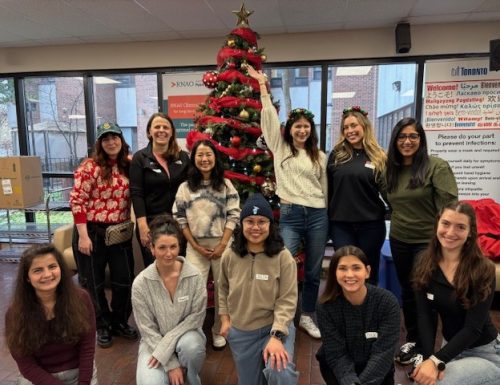 A group of people posing together indoors in front of a decorated Christmas tree with red ribbons and ornaments, topped with a gold star. The setting includes large windows in the background, a beige couch to the right, and a poster with text on the wall. The group is arranged in two rows, with some standing and others kneeling on the floor.