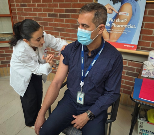 A member of the care team wearing a white lab coat administers a flu shot to a person sitting in a black chair. The person receiving the vaccine is wearing a dark blue shirt and has an ID badge hanging around their neck. In the background, there is a brick wall with a poster promoting flu shots, along with a table holding medical supplies.