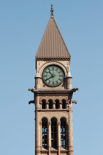 Close-up view of the Old City Hall clock tower in Toronto against a clear blue sky. The tower features a large round clock with Roman numerals, set in a decorative frame.