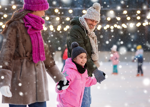 Two adults and a child skating on a snowy outdoor rink covered by string lights