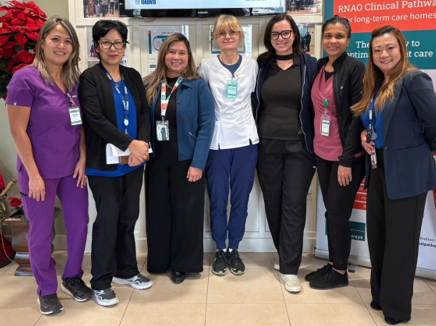 The Minister of Long-Term Care stands with 5 people together indoors in front of informational posters and a wall-mounted screen. They are wearing professional attire, including scrubs and ID badges.