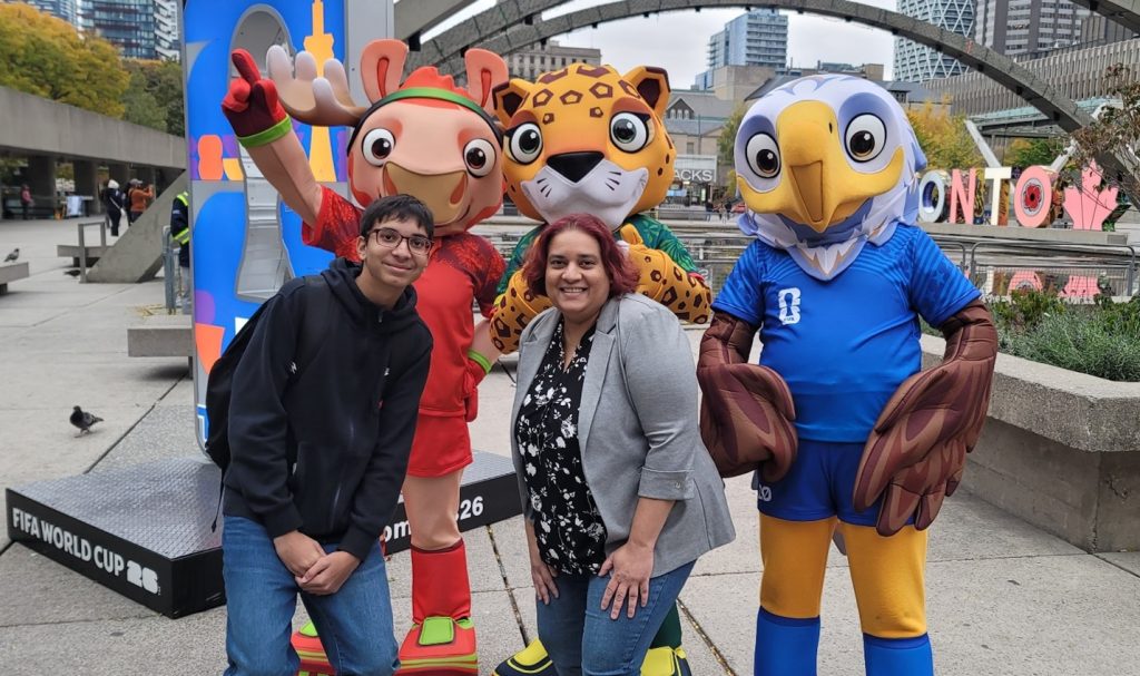 Three colourful mascots pose outdoors in front of the Toronto sign at Nathan Phillips Square. The mascots include a moose in a red outfit, a jaguar in green, and an eagle in a blue jersey. Two people stand between the mascots, and tall buildings and autumn trees are visible in the background.