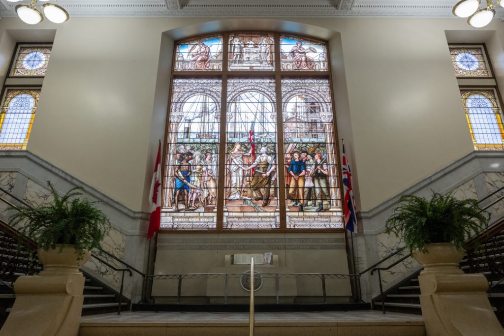 Large stained glass window at Old City Hall depicting a historical scene with figures in period clothing, flags, and architectural arches. The window is framed by marble walls and flanked by Canadian and British flags. Two staircases with dark railings lead up to the landing, and potted plants sit on pedestals on either side.