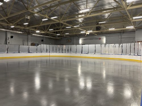 An indoor ice rink with a smooth, freshly prepared ice surface. The rink is surrounded by white boards with a yellow base trim and clear protective glass panels. Overhead, metal beams and bright fluorescent lights illuminate the space, and the ceiling has a reflective metallic finish. A sign with text is visible on the far wall above the boards. 