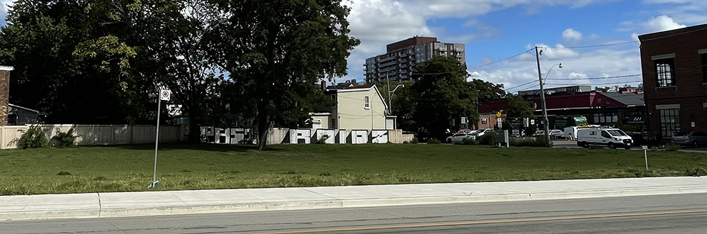 An open grassy lot with a few mature trees, surrounded by houses, sidewalks, and nearby commercial buildings.