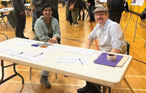 Two people sit at a table in a gymnasium during a public consultation event, with forms, pens and materials spread out in front of them and other attendees gathered in the background.
