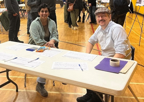 Two people sit at a table in a gymnasium during a public consultation event, with forms, pens and materials spread out in front of them and other attendees gathered in the background.