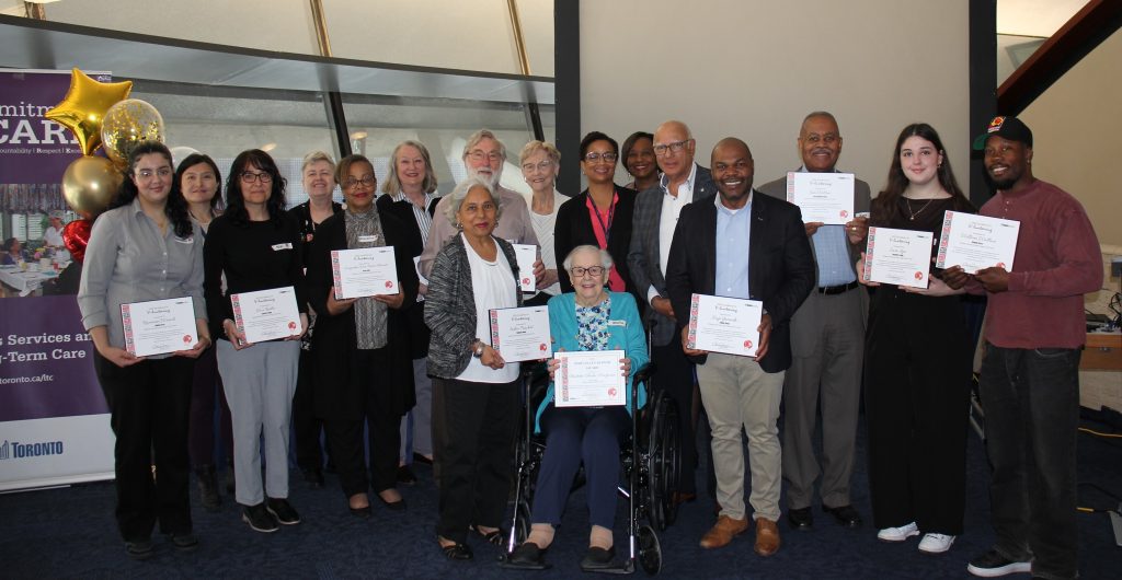 A large group of volunteers pose together for a photo at an awards event, each holding a certificate, with balloons and an event banner displayed to the side.