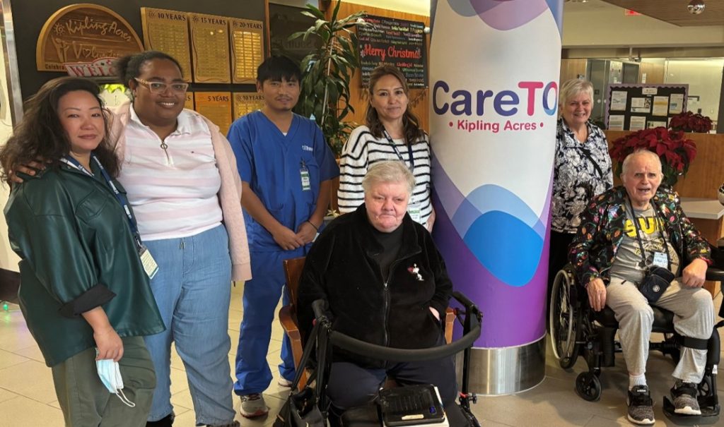 A group of residents and staff gathered in the lobby at Kipling Acres beside a CareTO display banner, with two people seated in wheelchairs and others standing behind them.