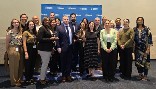 A large group of staff stand together for a photo while celebrating receiving the City Manager’s Innovation Award, with several people holding trophies in front of a blue City of Toronto backdrop.