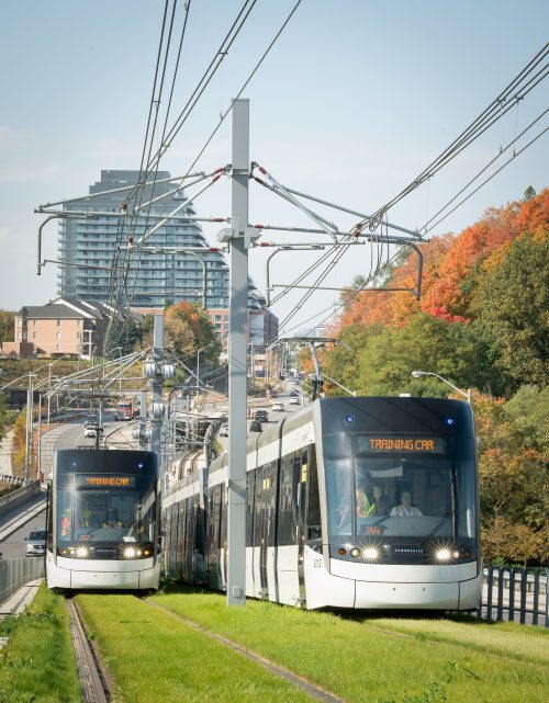Elginton LRT - Vehicles shown driving side by side with Training Car written on the front. The vehicles are going uphill along Eglinton on a sunny day.