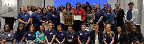 A large group of SSLTC staff pose together for a group photo, with two people in the front holding a large wooden plaque and a certificate.