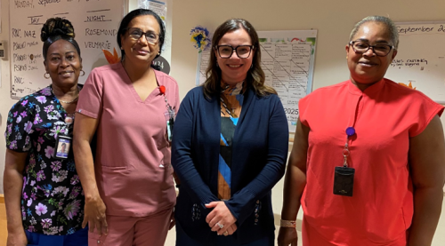 Minister of Long-Term Care and three long‑term care staff stand together in a hallway in front of a whiteboard with schedules and notices posted behind them.