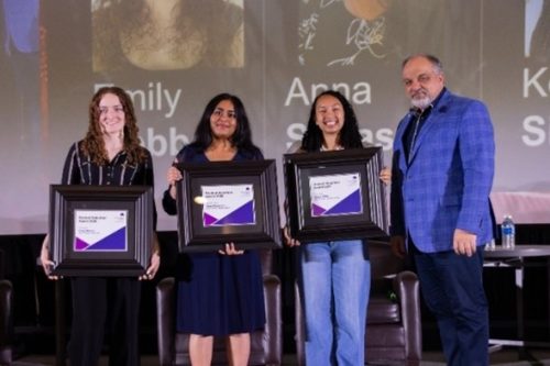 Three people stand on stage holding framed Student Volunteer Awards, with a fourth person standing beside them in front of a large projected backdrop.