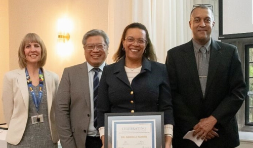 Four people stand together at an awards event, with the person in front holding a framed certificate while posed for a group photo indoors.