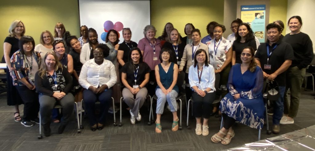 A large group of staff stand and sit together for a group photo in a meeting room, with balloons and a presentation screen in the background.