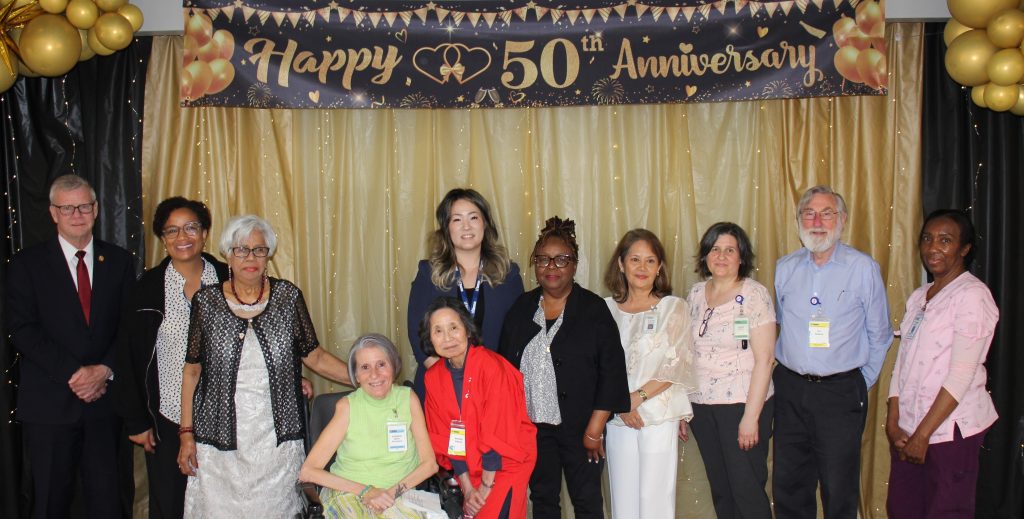 A group of people, including MPP John Jordan, staff and residents, pose together in front of a black and gold backdrop with a banner that reads ‘Happy 50th Anniversary.’ Gold balloons decorate the sides, and one person is seated in a wheelchair while others stand behind them.