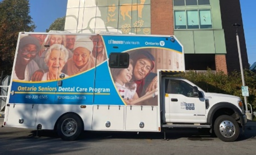 A mobile dental clinic truck for the Ontario Seniors Dental Care Program is parked outside a building, featuring large images of seniors and program branding on the side.