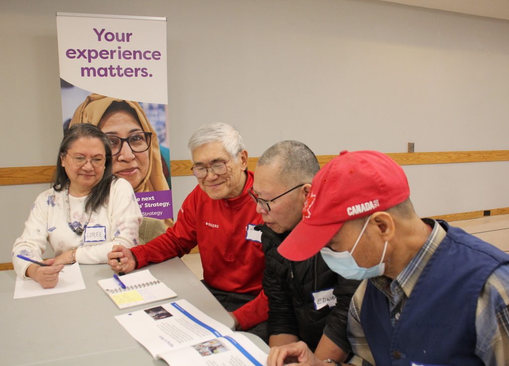 A group of four participants sit together at a table during a workshop, looking at printed materials and taking notes, with a banner in the background that reads ‘Your experience matters.’