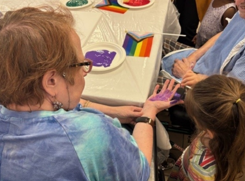 A resident sits at a table while a child helps apply purple paint to their hand for an art activity. Paint trays and a small Pride flag are on the table.