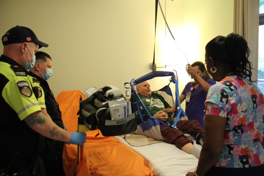Paramedics and long‑term care staff assist a resident using a mechanical lift in a bedroom during a safe‑lifting procedure.