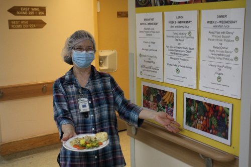 A staff member wearing a hairnet stands in a hallway holding a plate of food, next to a wall display showing the weekly long‑term care menu.