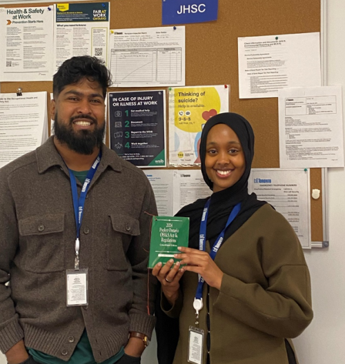 Two staff members stand in front of a Joint Health and Safety Committee bulletin board, with one person holding a green health and safety guidebook.