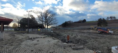 Wide view of a construction site with excavated ground and construction equipment, including an excavator and skid-steer, on the right. A paved area with removed bricks is in the foreground, a leafless tree stands near the center, and a small building and light poles are on the left under a partly cloudy sky.