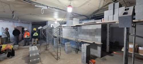 Interior construction scene with workers wearing safety helmets and high-visibility vests beside metal scaffolding. Concrete block walls are partially built, with stacks of cinder blocks and tools on the floor and temporary lighting illuminating the space.