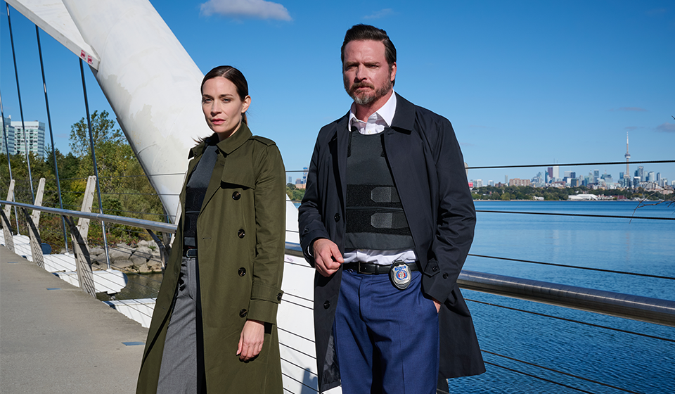 Two people standing on a bridge with the Toronto skyline in the background.