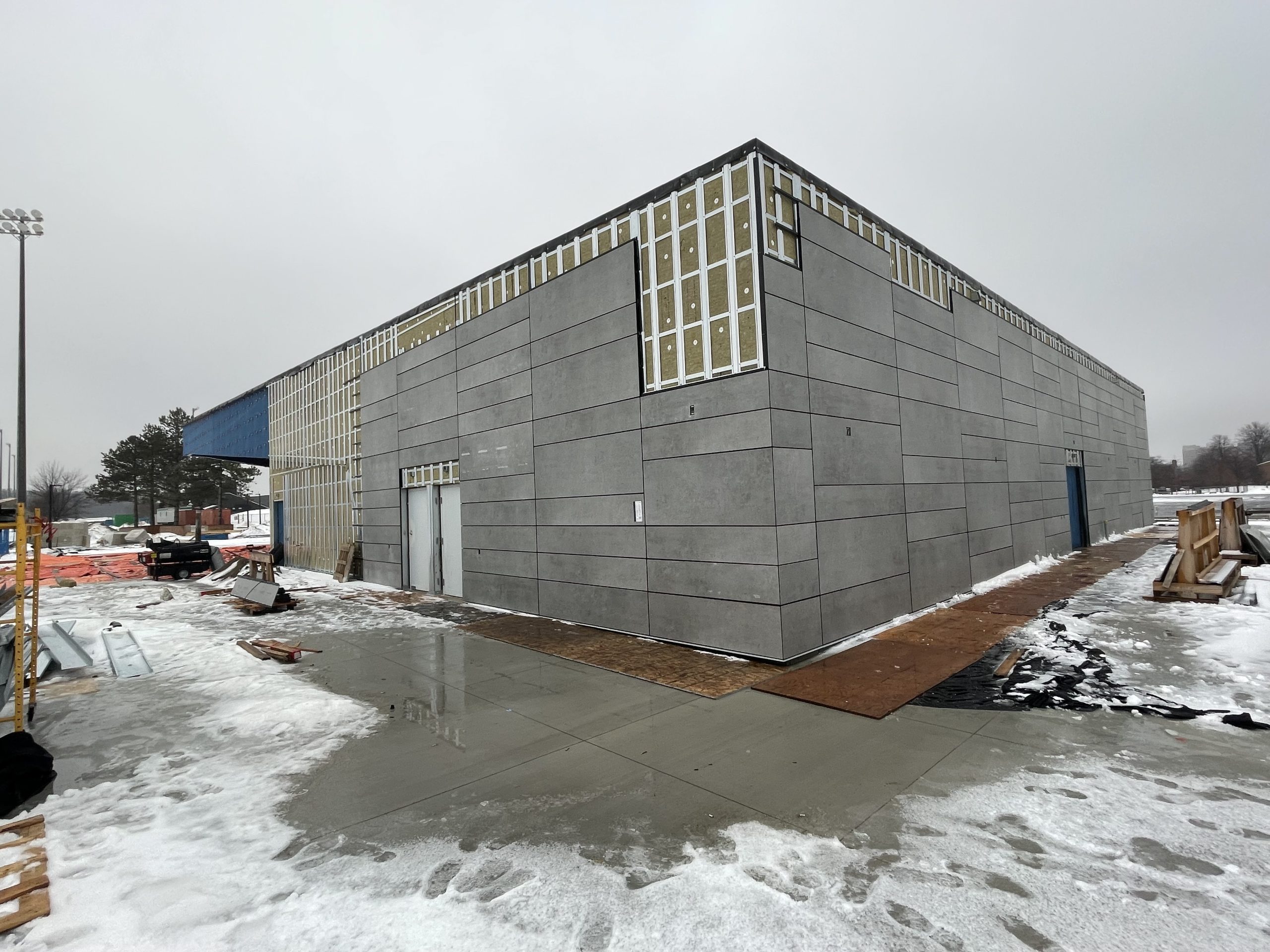 A large rectangular building under construction with gray cladding panels partially installed. The upper section shows exposed insulation and framing. The ground around the building is wet with patches of snow and ice, and construction materials are scattered nearby. The sky is overcast, creating a cold, wintery atmosphere.