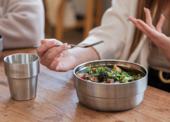 A person holding a fork at a table with a reusable steel bowl containing a colourful salad and a reusable steel cup.