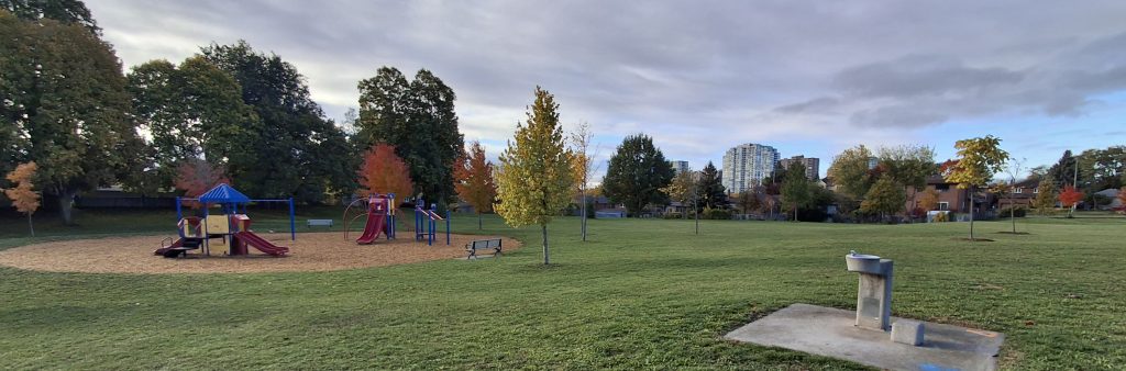 A large open park with green grass, a playground with slides and climbing structures on a sand surface to the left, and a drinking fountain on a concrete pad in the foreground. Scattered trees with autumn colours surround the park, and mid rise buildings are visible in the distance under a cloudy sky.