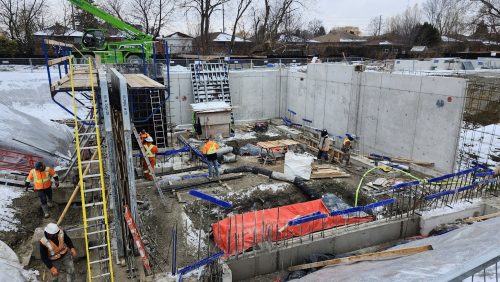 A wide view of a snow covered construction site. A long green stormwater tank structure is being installed on the left side, with machinery, materials, and dirt piles spread throughout the area. Trees and houses line the background under a cloudy sky.
