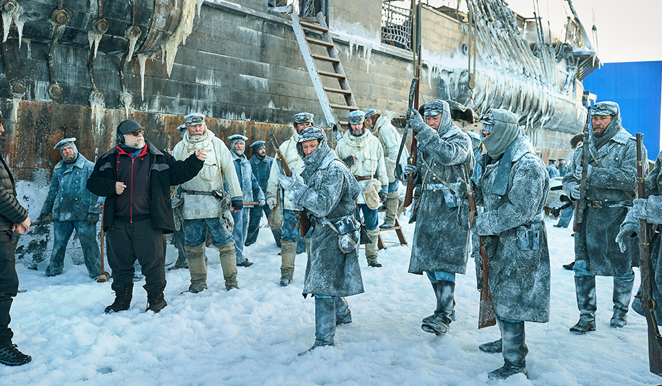 Actors on set of an old ship in winter time dressed in costumes holding muskets with a director point at something.