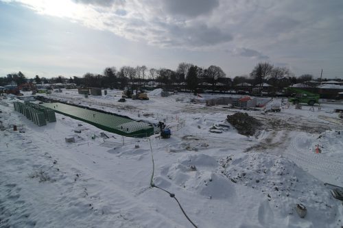 A construction site in winter showing workers inside a partially built concrete foundation. Several people in safety gear are installing and preparing formwork along the walls. A ladder and elevated platform are positioned on the left, with equipment and materials scattered across the snowy ground.