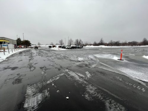 A newly paved parking lot covered with slushy snow and ice patches. Several vehicles are parked in the distance near snowbanks, and a single orange traffic cone stands on the right side. The background includes bare trees and an overcast gray sky, indicating winter conditions.