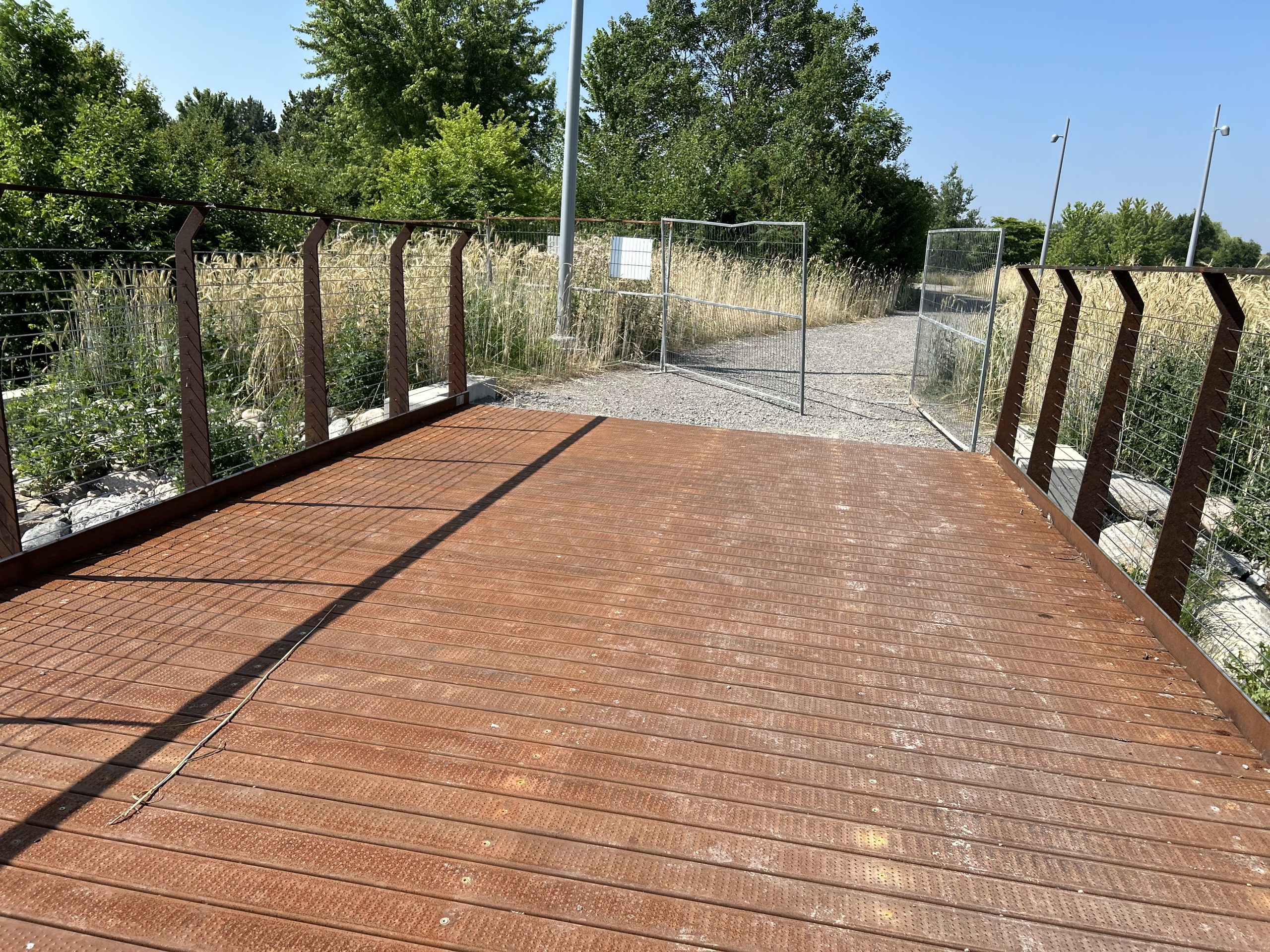 Ground-level view of a section of the boardwalk under installation along a gravel path. The walking surface is made of steel planks. On either side are railing frames with cables. Tall grasses and trees surround the area under a blue sky.