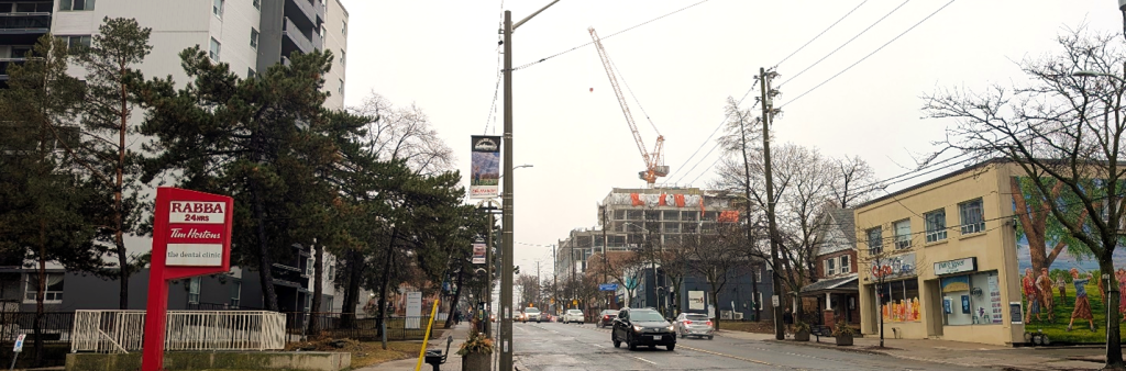Street view image of Dundas Street West facing west. The image includes a mid-rise residential building and low-rise commercial retail buildings, as well as a building under construction in the distance.
