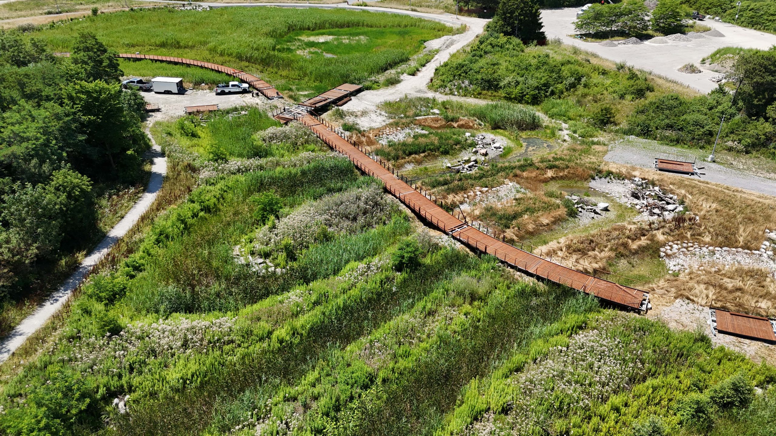 Aerial view of a long boardwalk under construction in a marsh setting filled with plants and trees. Construction materials and vehicles are visible on gravelled areas surrounding the boardwalk.