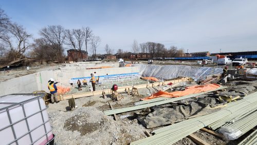 A wide view of an active construction site showing workers wearing safety vests standing inside a large excavated area with concrete foundation walls. The centre of the site includes forming materials and rebar for a pool basin, with protective tarps and equipment scattered around. Trees and buildings are visible in the background under a clear sky.