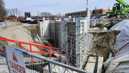 A construction area with exposed concrete foundation walls, rebar, and soil excavation. A temporary metal fence with a “Danger Excavation” sign is in the foreground. Construction machinery, materials, and a large green lift are visible across the site, with buildings and trees in the background.