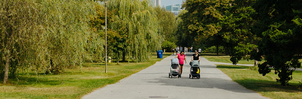Two people walking down a wide paved path in a tree lined park with the CN Tower in the background on a sunny day.