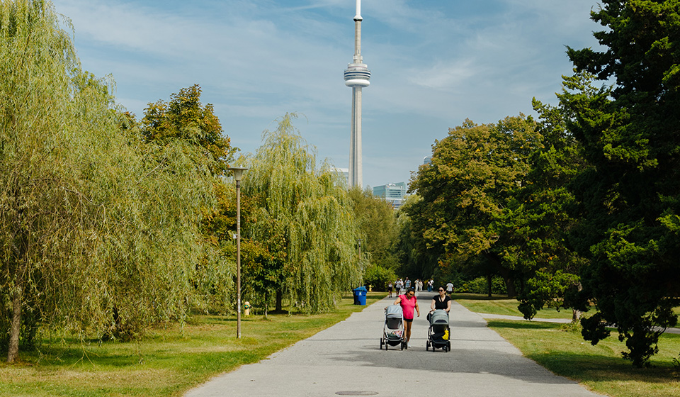 Two people walking down a wide paved path in a tree lined park with the CN Tower in the background on a sunny day.