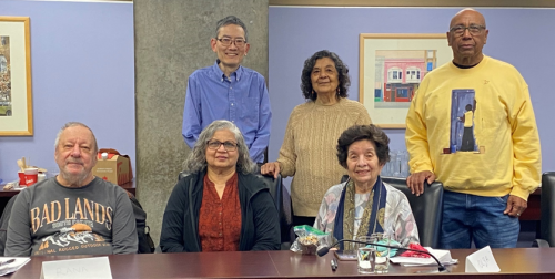 Six adults pose behind and around a conference table in a meeting room, with framed artwork on the wall and papers, notebooks, and glasses on the table. Three people are sitting, and three people are standing behind them.